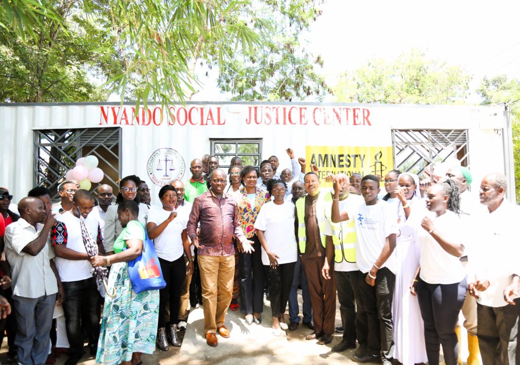 A group of people smiling on a sunny day behind a metal container with words writtern Nyando Justice Centre